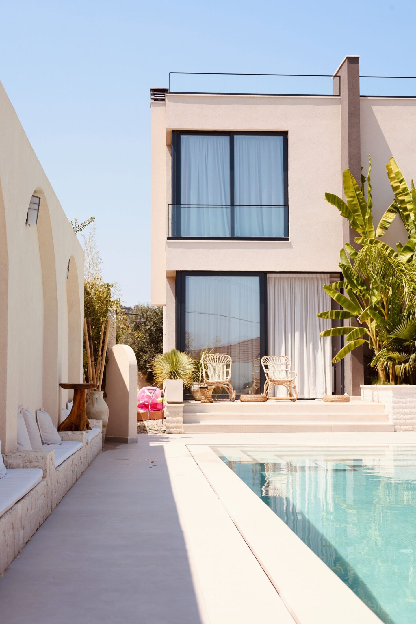 The pool runs the length of the villa's cream stucco facade, with banana palms and rattan chairs on the terrace.